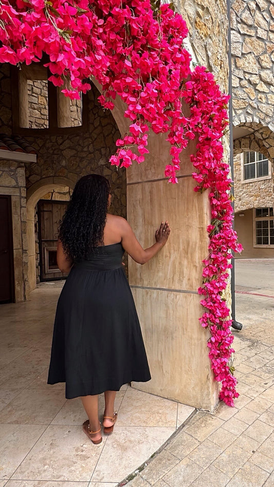 back view of a woman in a flowy black midi dress standing next to a stone wall with draped pink flowers