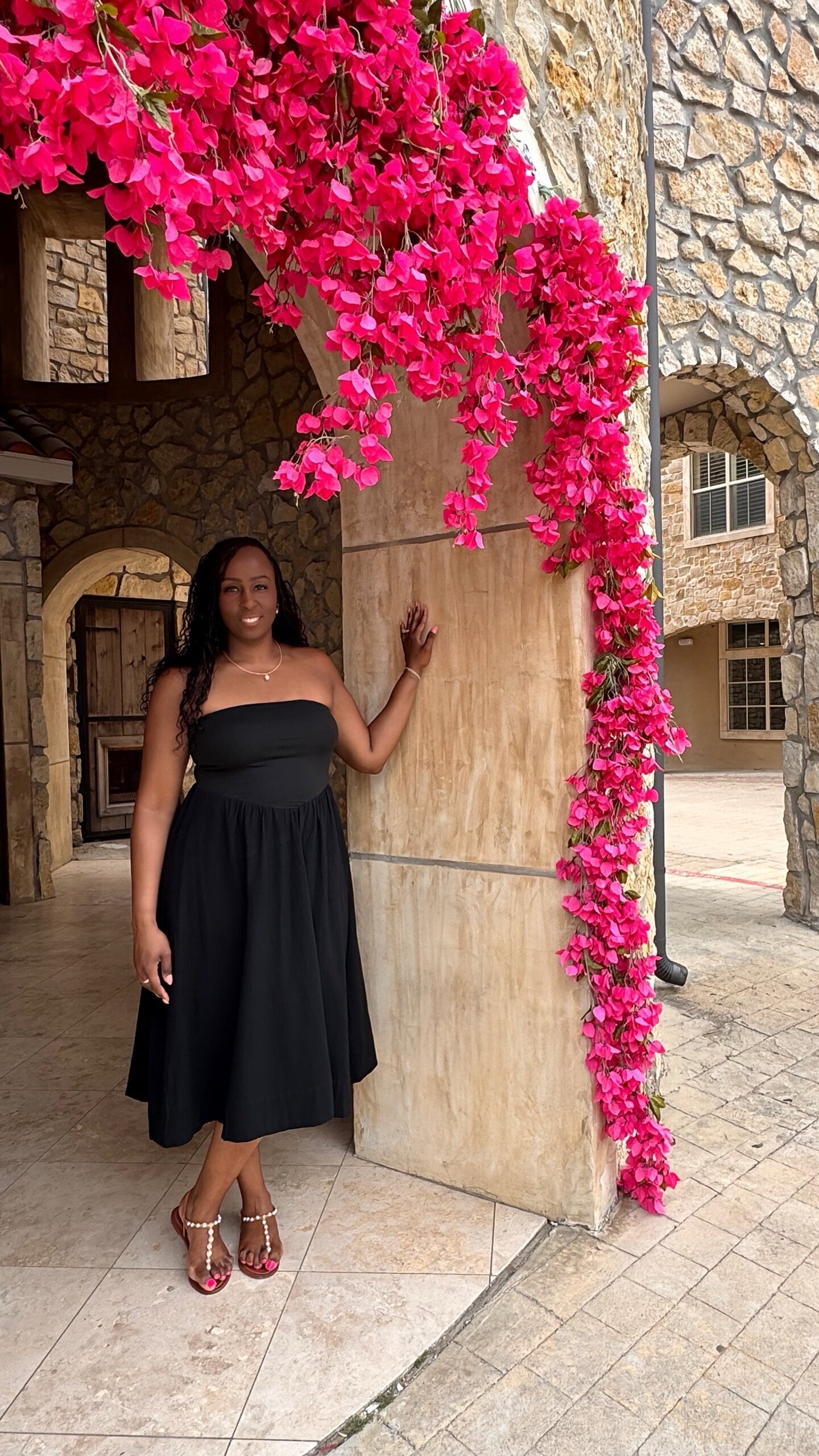 a woman in a flowy strapless midi black dress standing next to a stone wall with pink flowers