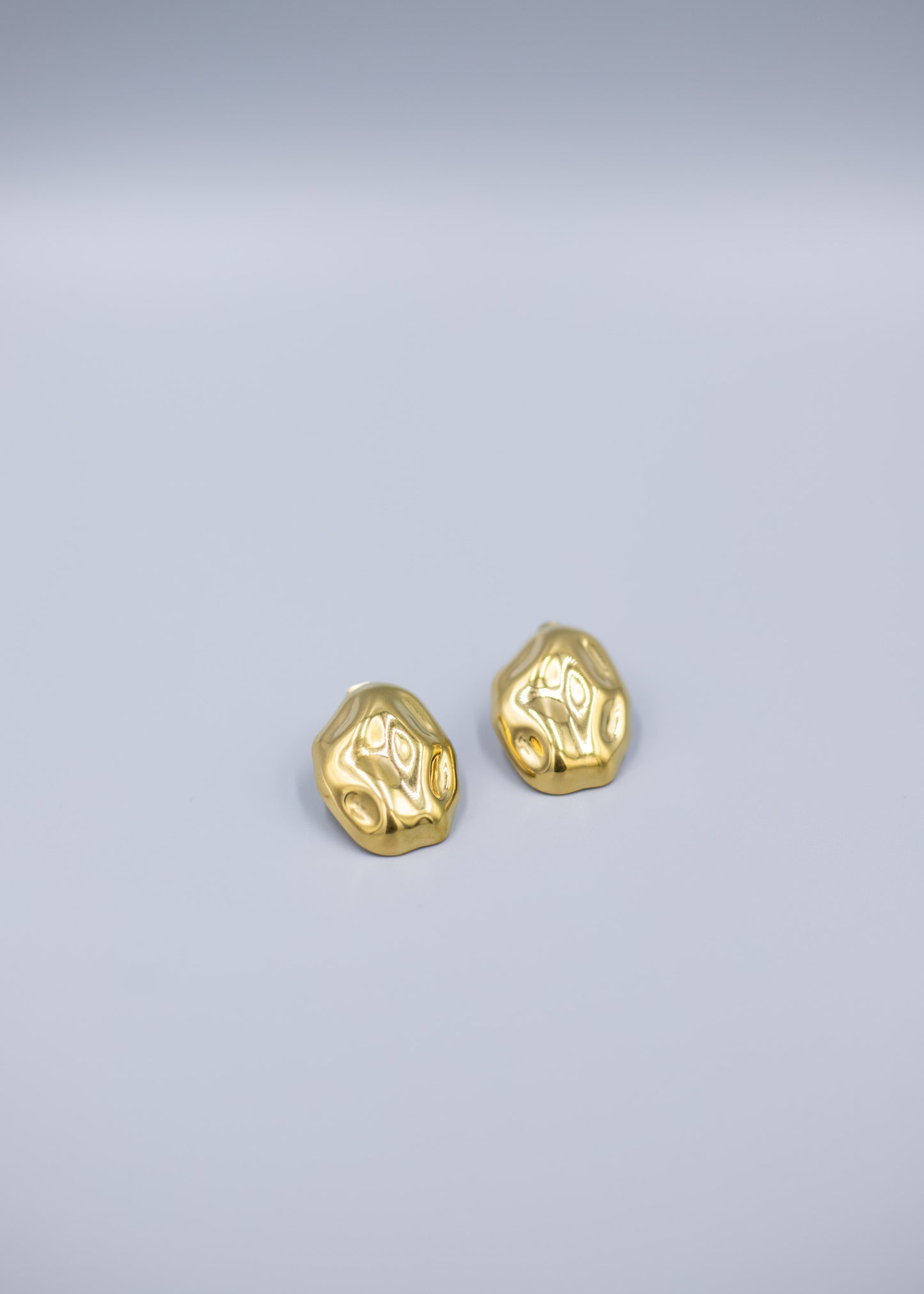 Angled view of a pair of 18k gold plated hammered stud earrings on a light gray background
