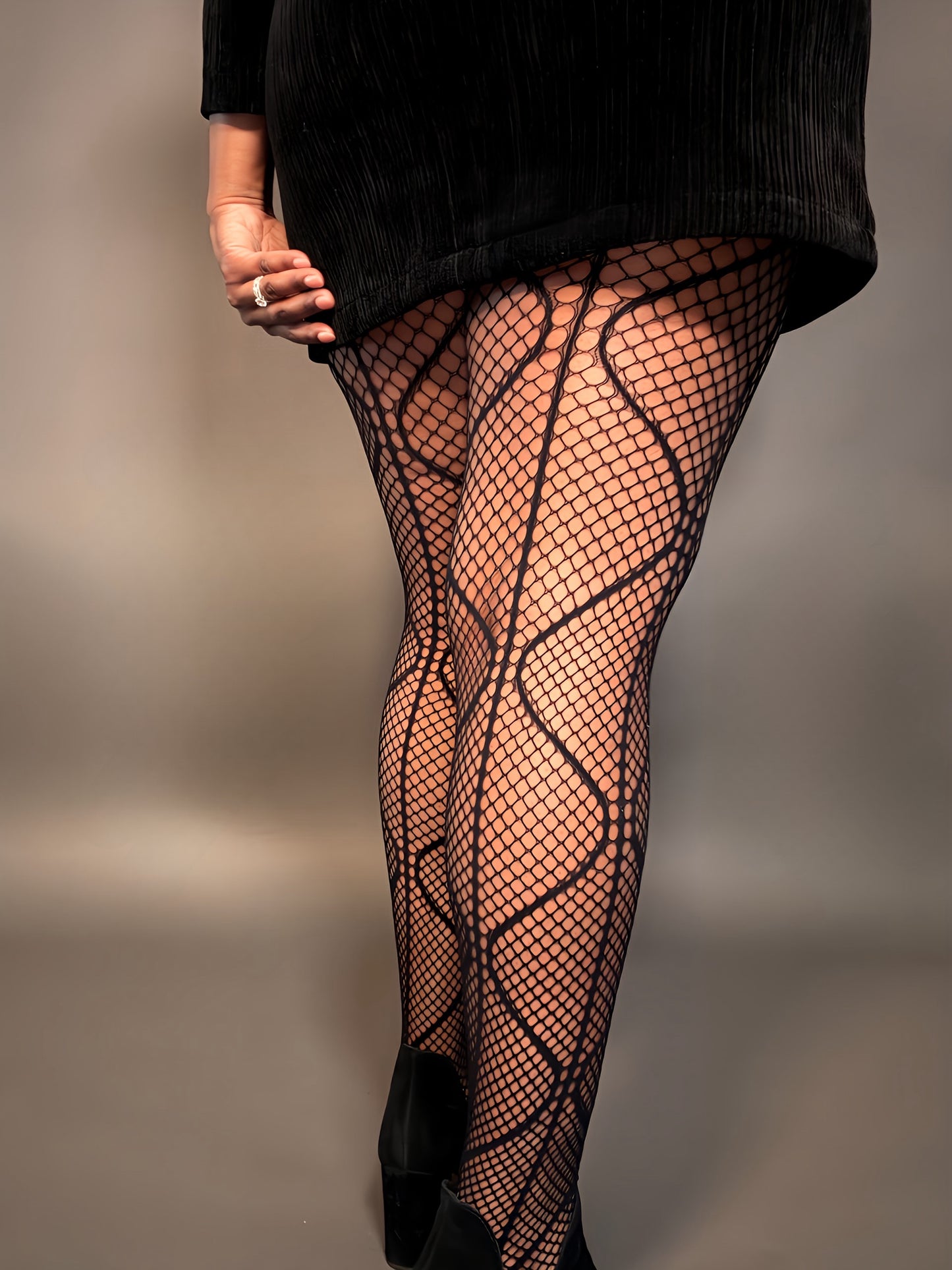 Detailed back view of elegant woman modeling Magnetic Waves Fishnet Tights, plus size hosiery for women, styled with a black dress and boots for a chic, trendy look.