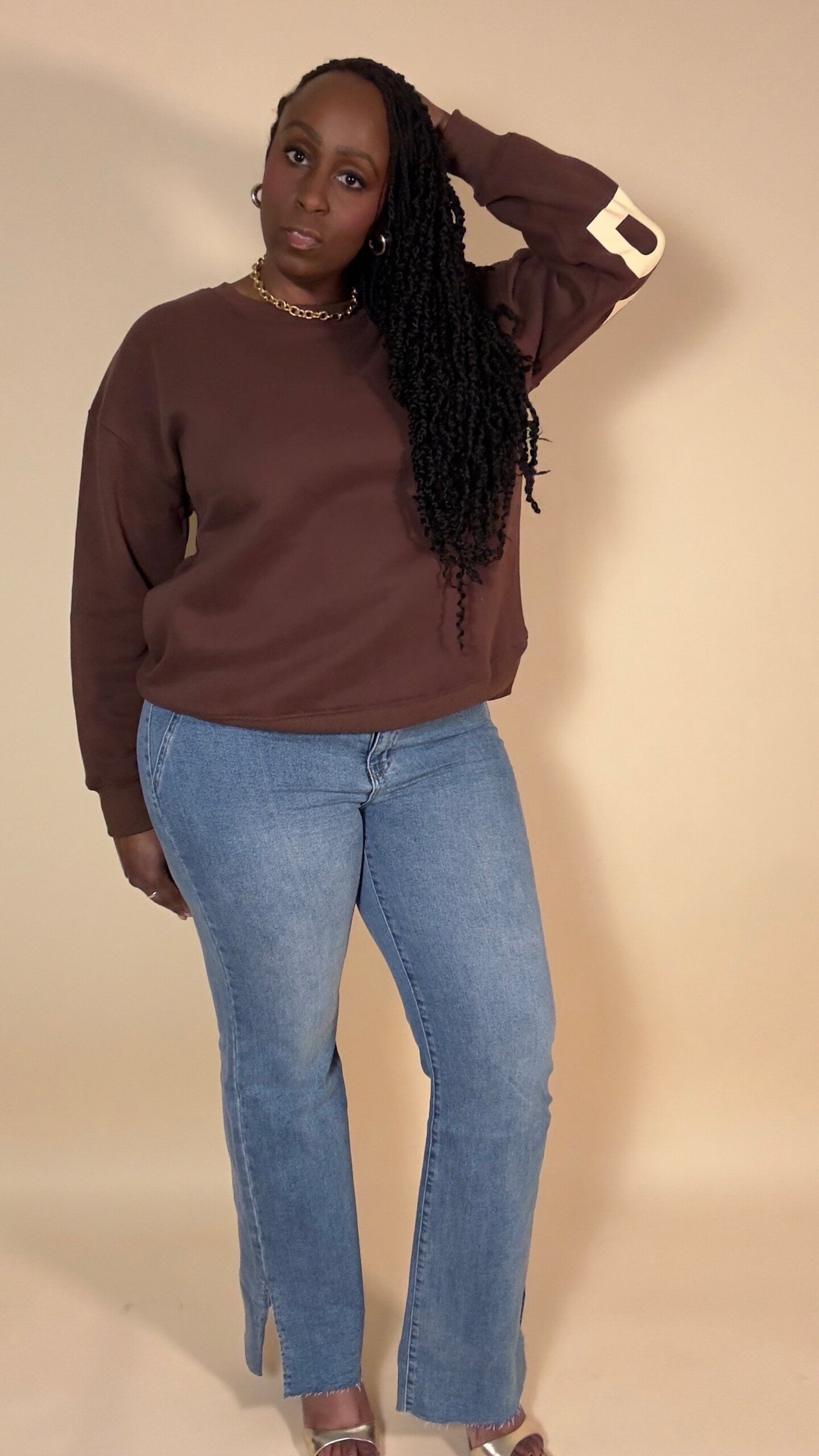Woman wearing a "BE YOURSELF Love yourself" brown sweatshirt and blue jeans against a beige background
