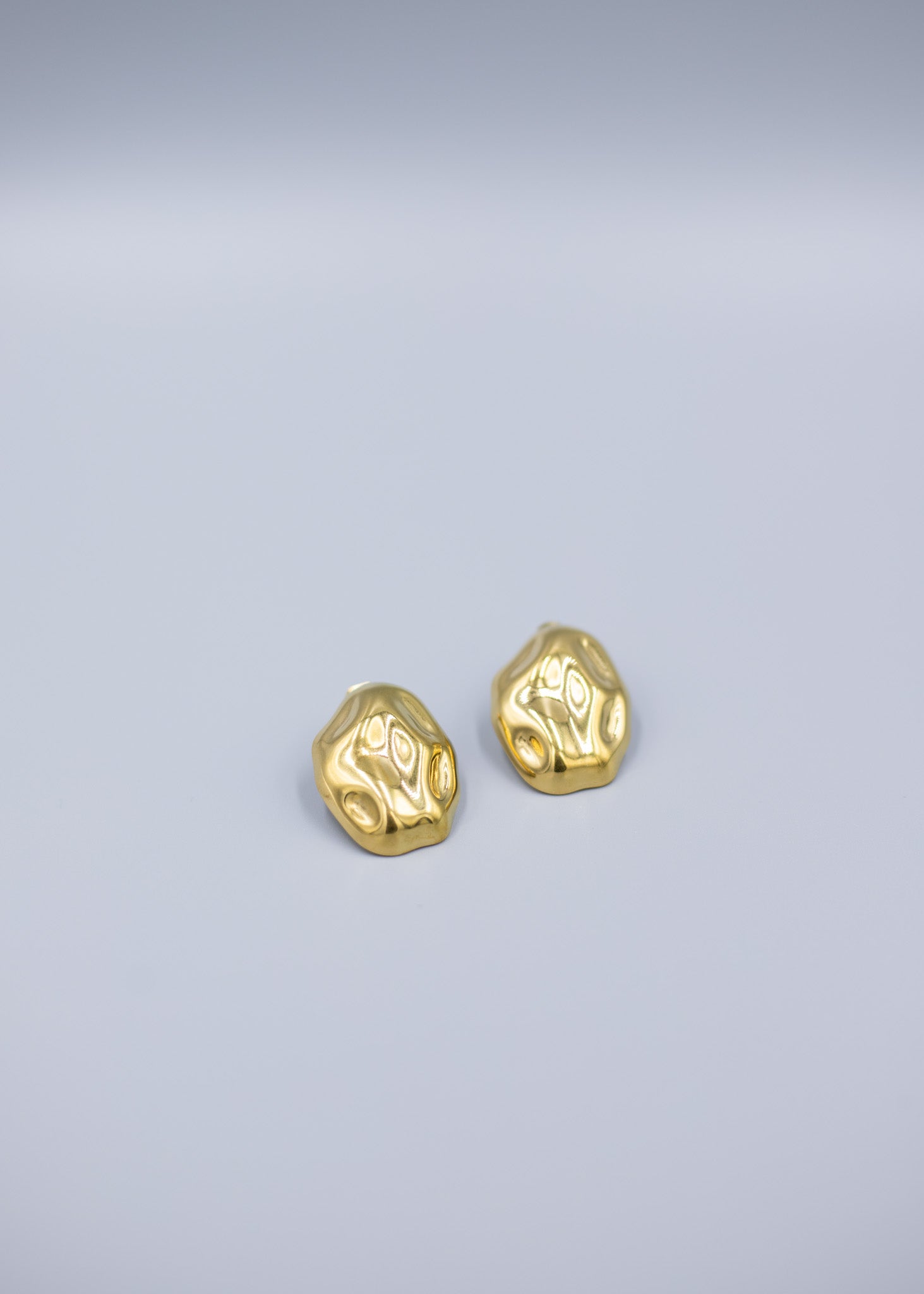 Angled view of a pair of 18k gold plated hammered stud earrings on a light gray background