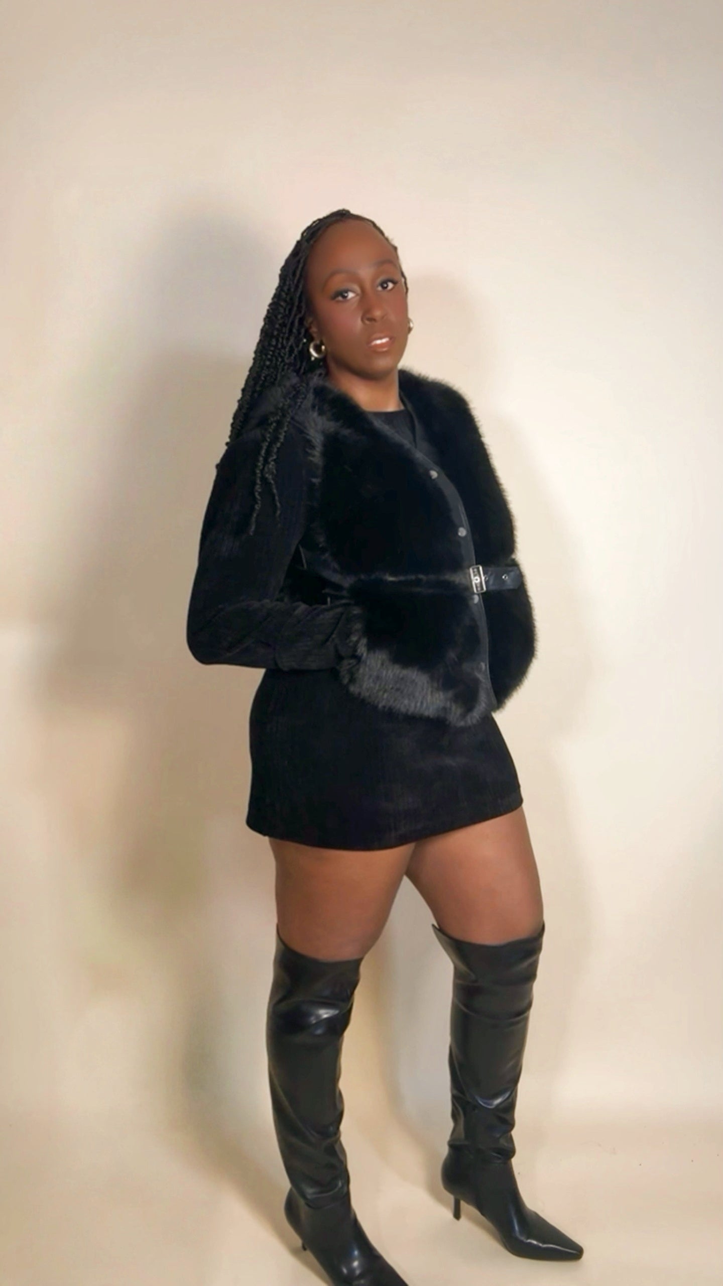 Person wearing a black fur vest and high-heeled boots on a plain background