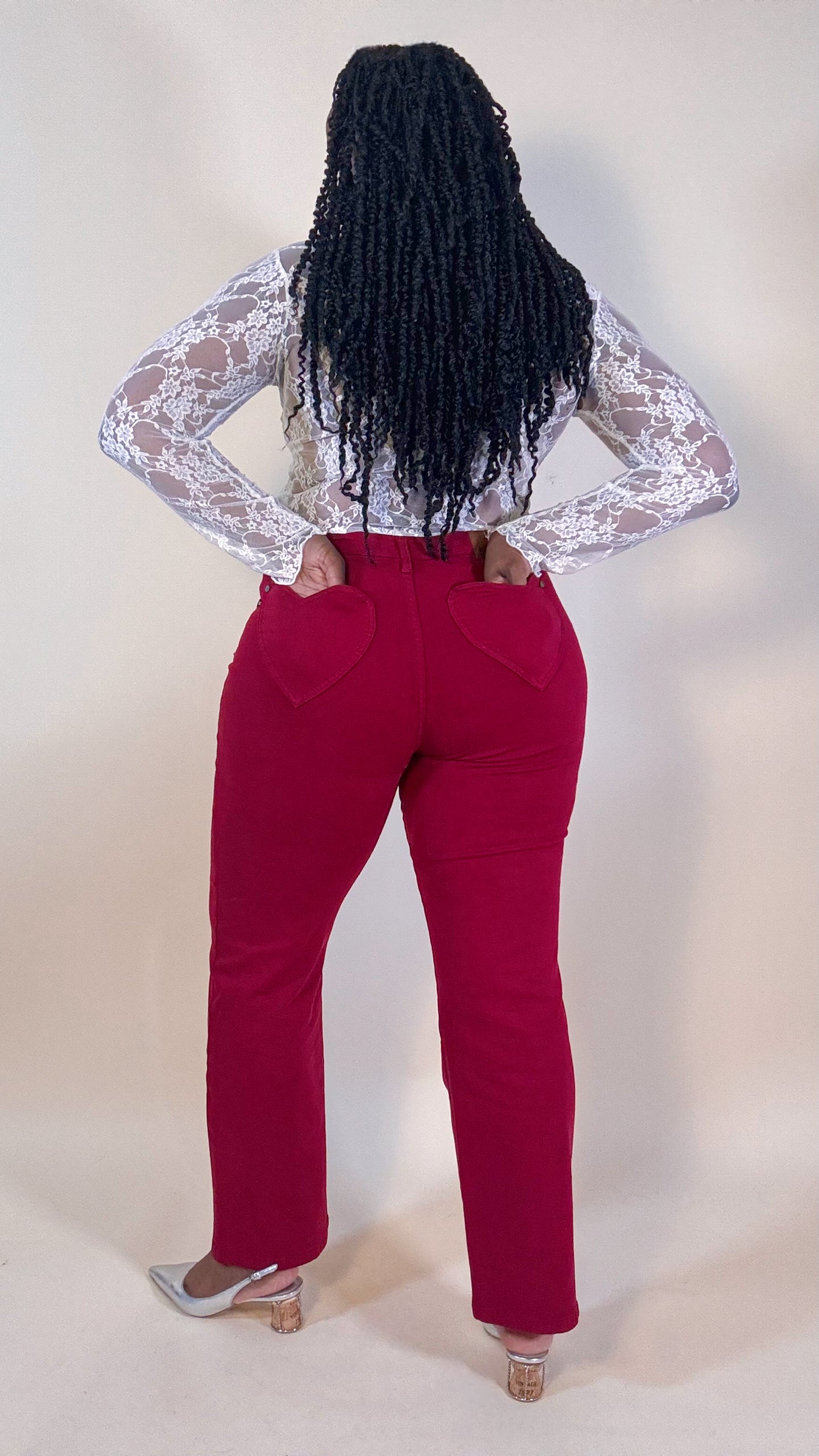 Woman wearing red jeans with heart pockets and a white lace top on a neutral background