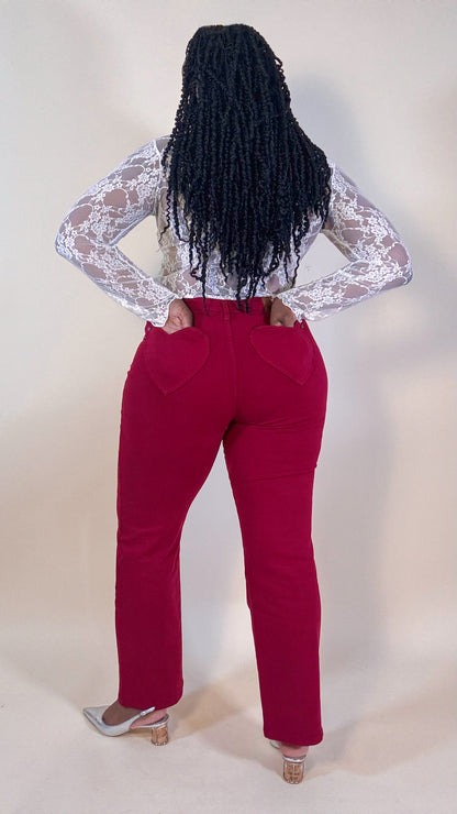 Woman wearing red jeans with heart pockets and a white lace top on a neutral background