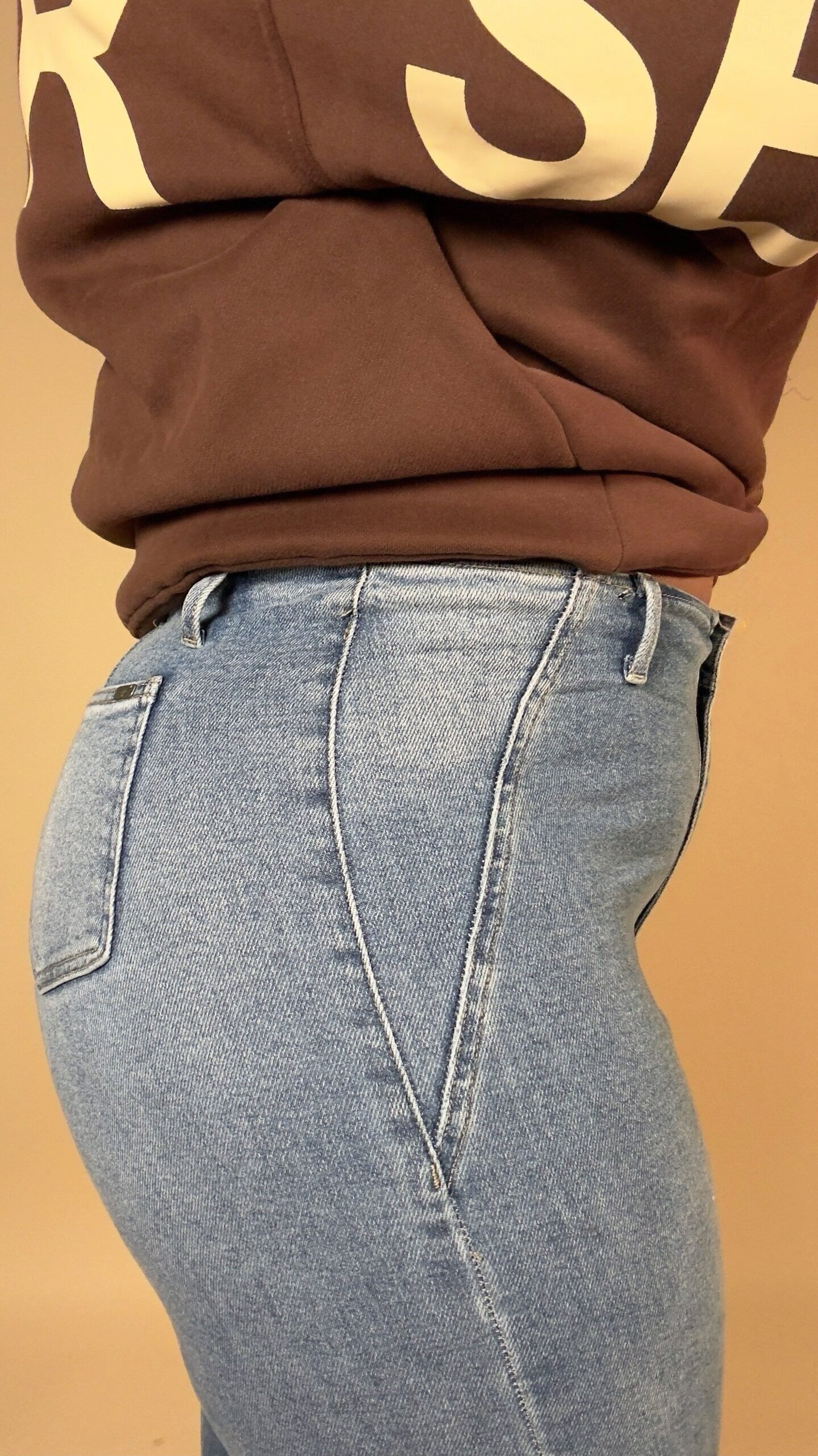 Person wearing a brown sweatshirt with "be yourself love yourself" text in beige on a beige background and light blue stretchy jeans on a beige background