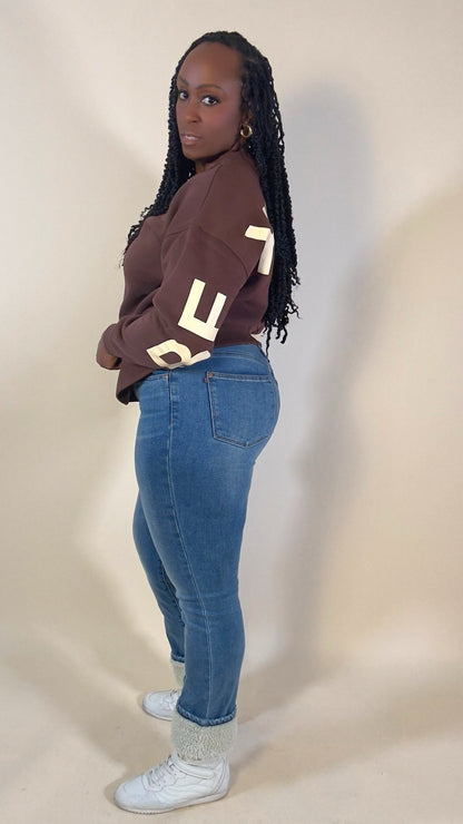 Person wearing a brown sweatshirt with large white letters and blue jeans with a Sherpa text cuff on a beige background