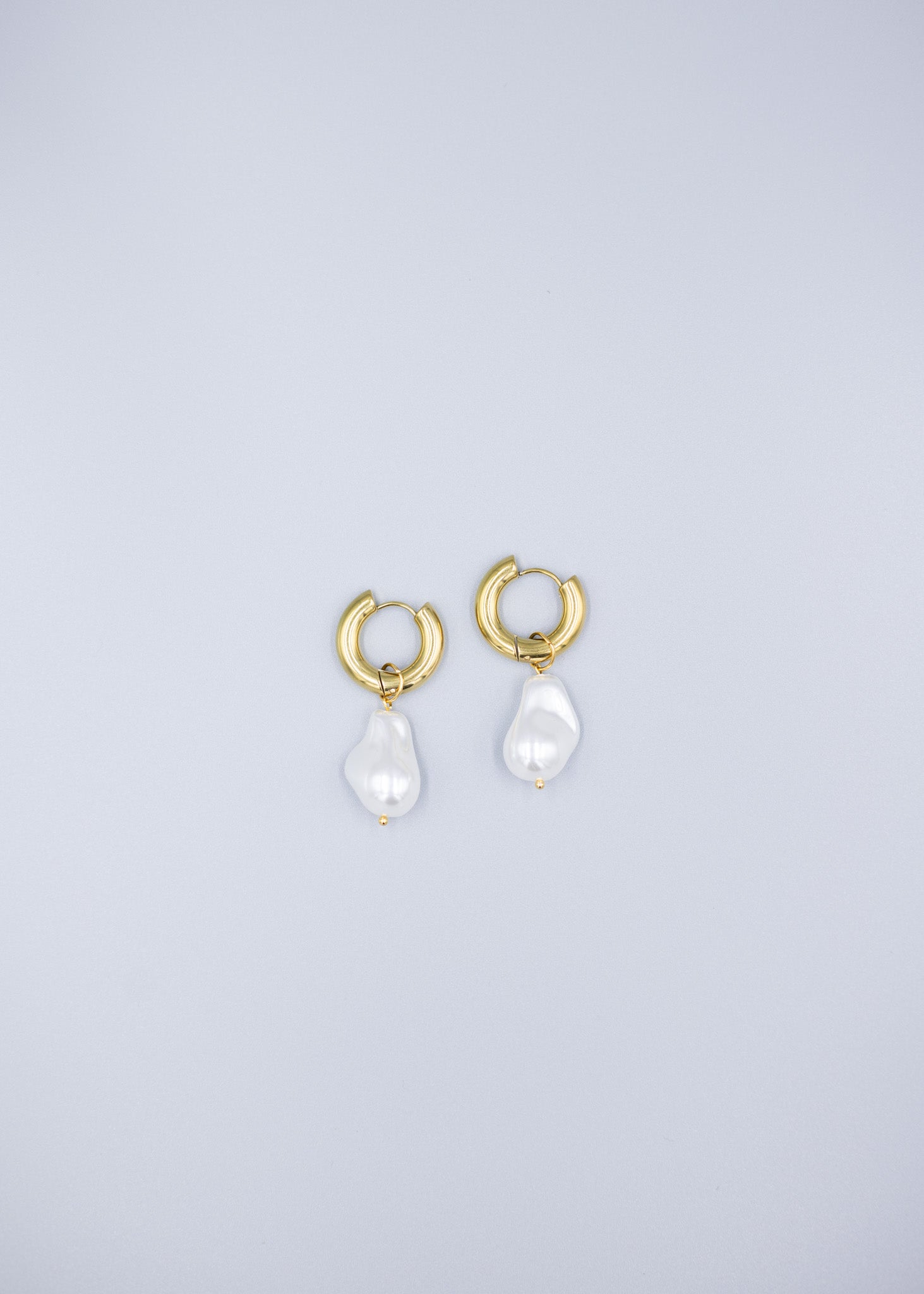 Distant view of a pair of 18k gold plated hoop earrings with removable dropped baroque pearls laid flat on a light gray background