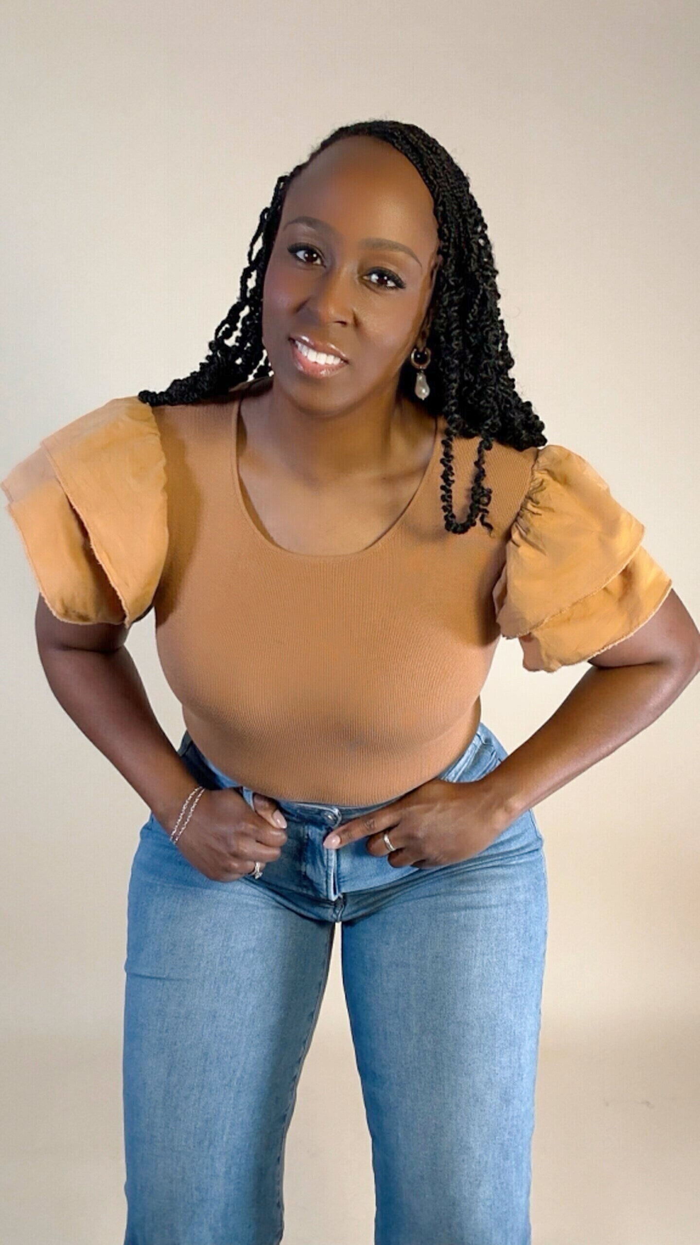 Woman wearing a light brown puff ruffle sleeve top and blue jeans against a plain background