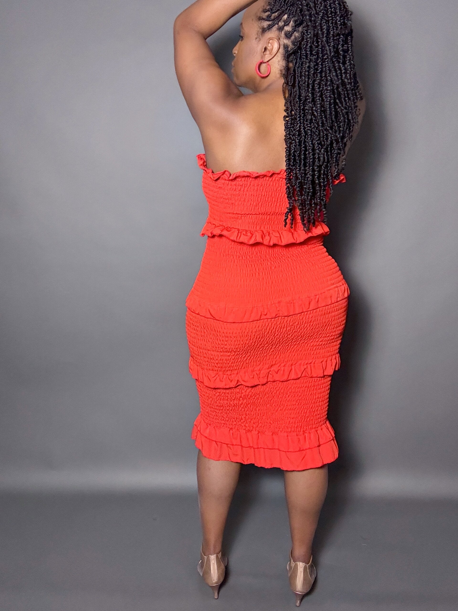 Woman wearing a red smocked shirred midi tube dress with ruffled details against a gray background