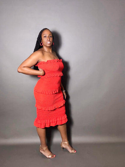 Woman wearing a red smocked strapless shirred midi tube dress with ruffled details against a plain gray background